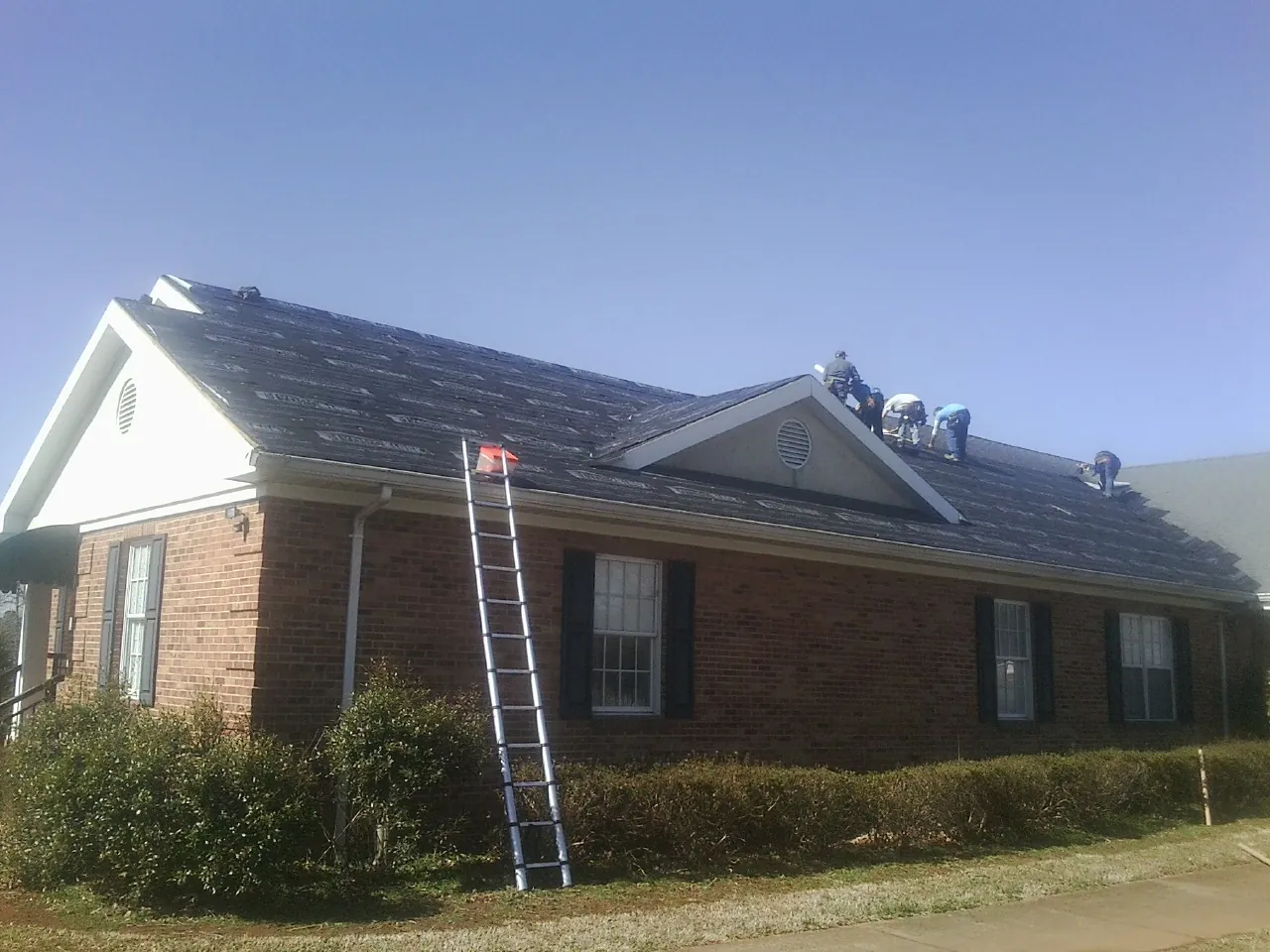 Crew tearing shingles off a brick ranch roof with a ladder set — Emergency Roof Repair work by our crew in Mission