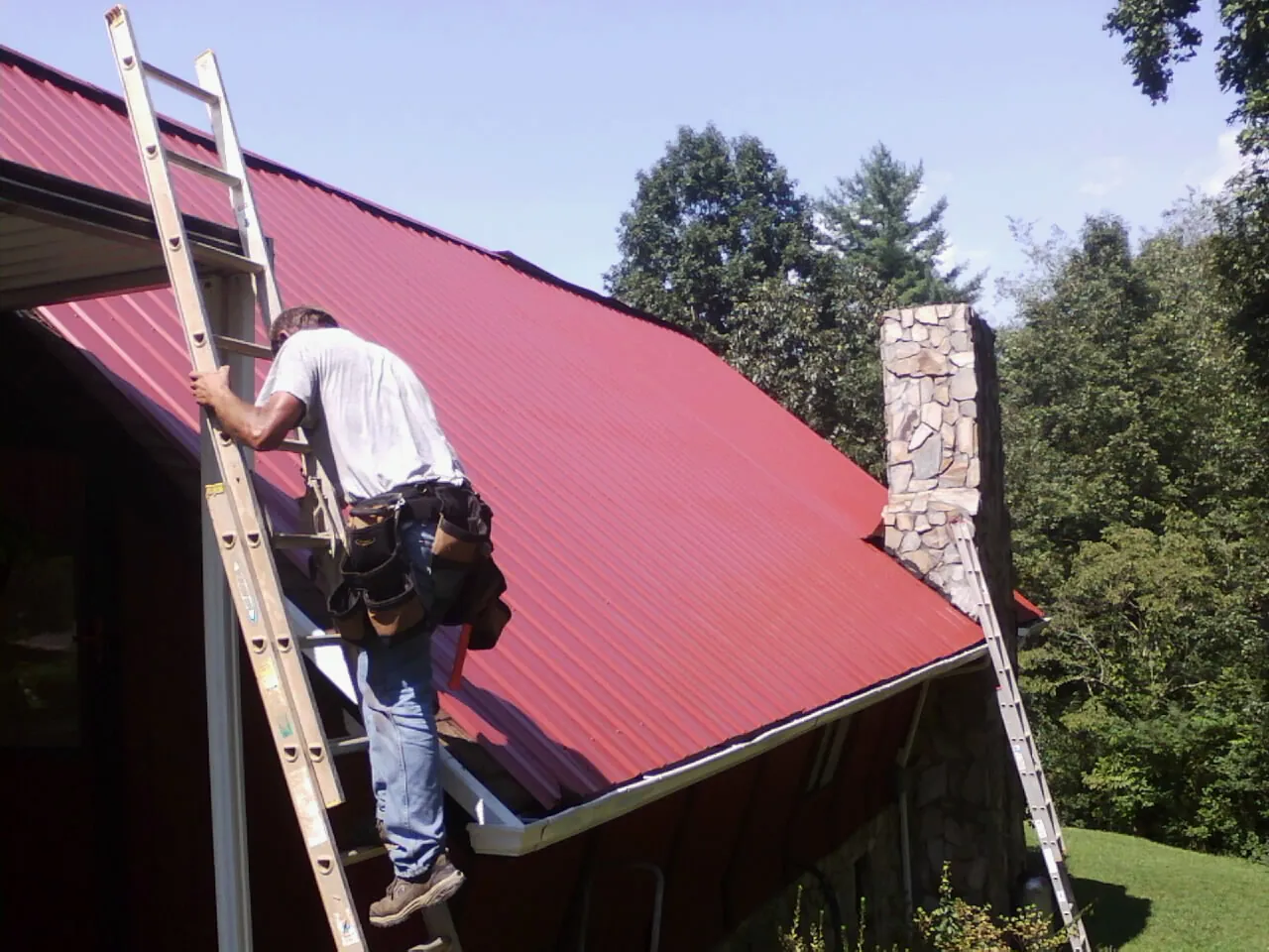 Roofer on a ladder beside a red metal roof and stone chimney — Soffit & Fascia Repair by Roofing Brothers in Mission